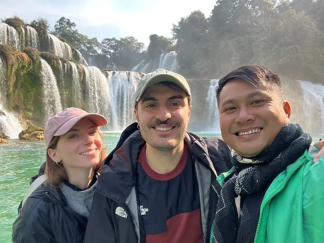 Three smiling friends taking a selfie with Ban Gioc Waterfall in background.