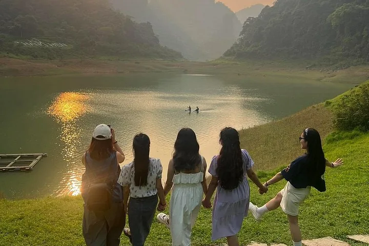 Five women holding hands by a serene lake at sunset, mountains behind.